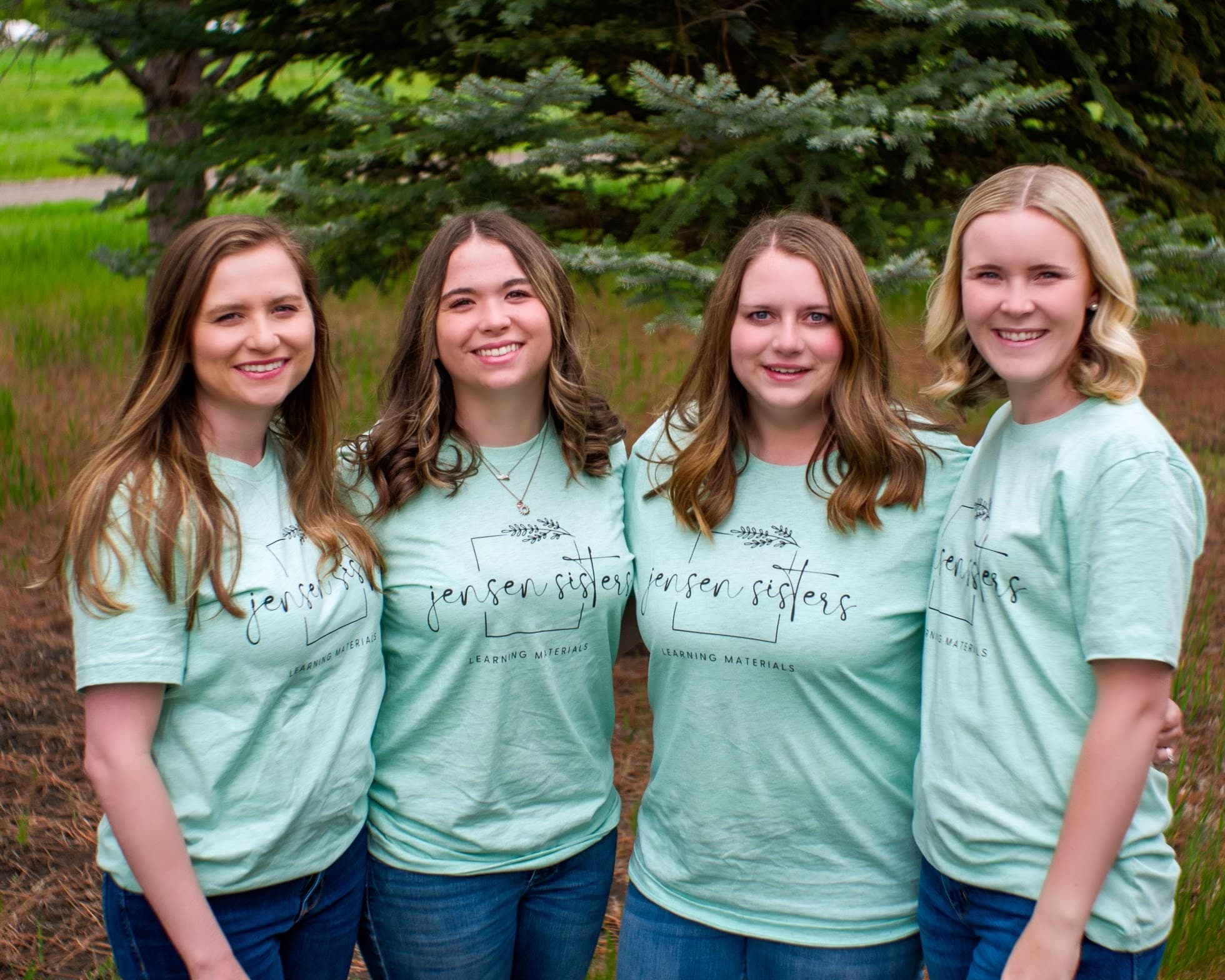 Janalie, Natalie, Kristle, and Ashlee — the four Jensen sisters, in matching mint Jensen Sisters tees in front of evergreens